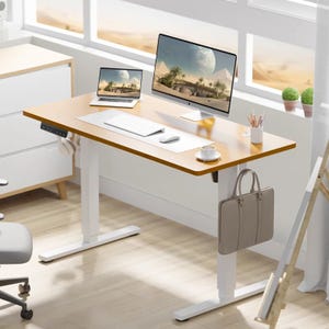 A modern home office setup with a laptop, a larger monitor, keyboard, mouse, coffee cup, stationery holder, and a bag hanging on the desk's side.