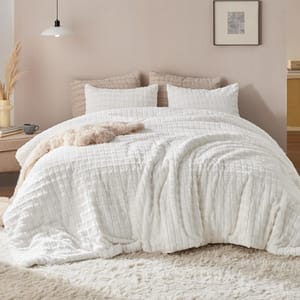 A cozy bedroom setup featuring a textured white quilted comforter, matching pillows, beige cushions, and a shaggy area rug underneath, accented by a framed artwork and minimalistic decor.