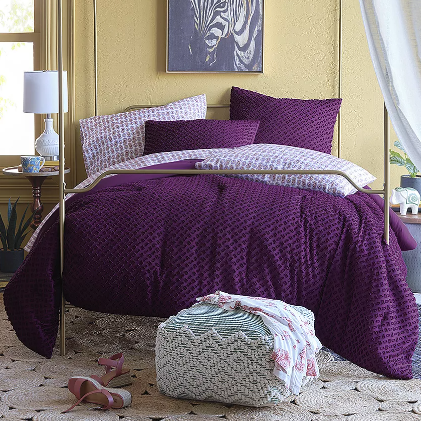A purple textured comforter set on a bed, accompanied by matching pillows, with a knitted pouf and sandals on the floor.