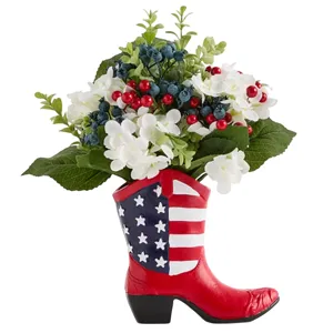 A patriotic-themed cowboy boot vase decorated with the American flag design, filled with artificial white flowers, red berries, and blue berries.