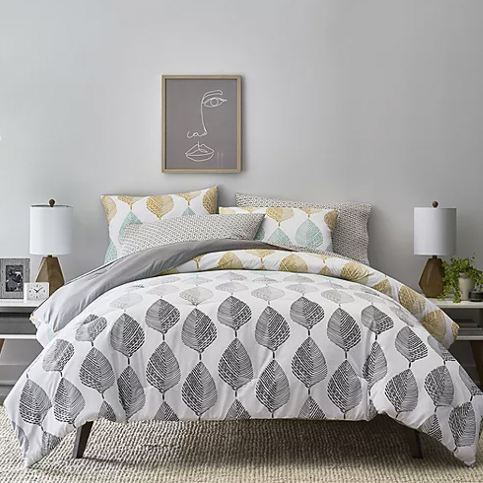 A bedding set with a leaf pattern, two white table lamps, and a framed abstract face artwork above the bed.