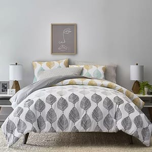 A bedding set with a leaf pattern, two white table lamps, and a framed abstract face artwork above the bed.