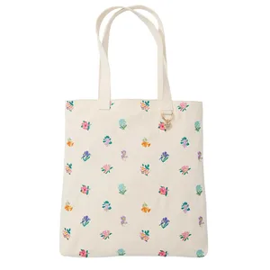 A cream-colored tote bag with a floral pattern featuring various colorful flowers. The bag includes dual handles and a small round charm attached to a ring near the top.