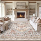 A cozy living room features a patterned area rug with warm tones, centered between two white sofas. The space includes a lit fireplace with a wooden mantle and minimalist decor on surrounding shelves.