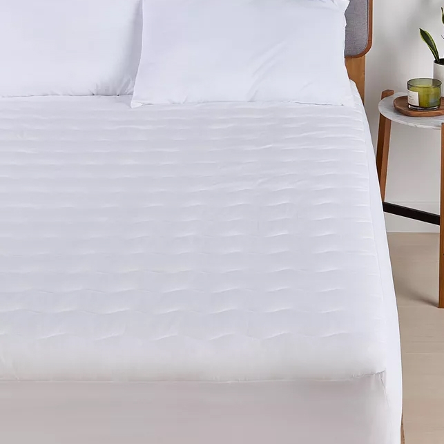A bed with a white quilted mattress protector and two pillows is shown, alongside a nightstand with a candle and a plant.