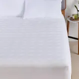 A bed with a white quilted mattress protector and two pillows is shown, alongside a nightstand with a candle and a plant.