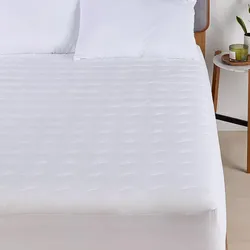 A bed with a white quilted mattress protector and two pillows is shown, alongside a nightstand with a candle and a plant.