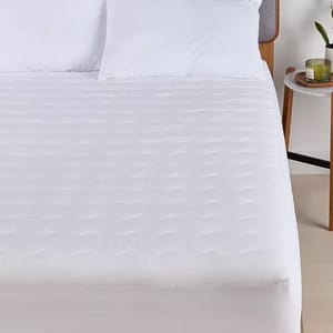 A bed with a white quilted mattress protector and two pillows is shown, alongside a nightstand with a candle and a plant.