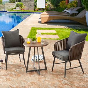 A 3-piece patio bistro set with two cushioned chairs and a round table. The chairs have dark cushions and woven backs, while the table holds a glass pitcher and glasses with orange juice. Set on a stone patio beside a pool.