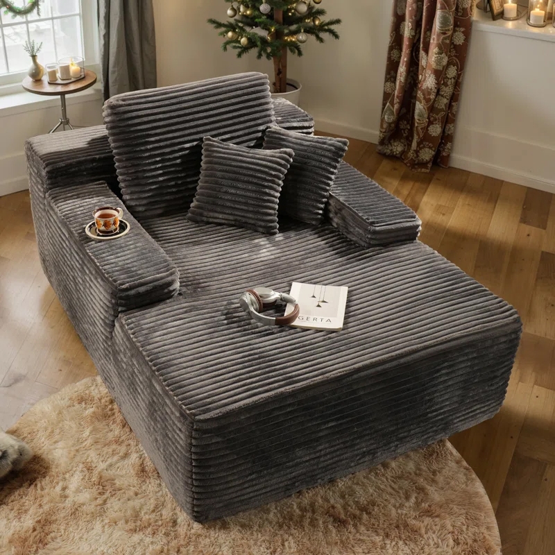 Gray corduroy oversized chaise lounge with two plush pillows, featuring wide armrests with cup holders, set in a cozy room with a wooden floor and soft rug, near a window and decorated Christmas tree.