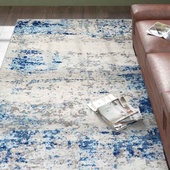 5' x 8' area rug featuring an abstract design with blue, gray, and white tones, providing a modern and artistic look. A brown leather sofa and a few magazines are visible as part of the room setting.