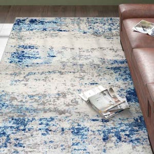 5' x 8' area rug featuring an abstract design with blue, gray, and white tones, providing a modern and artistic look. A brown leather sofa and a few magazines are visible as part of the room setting.
