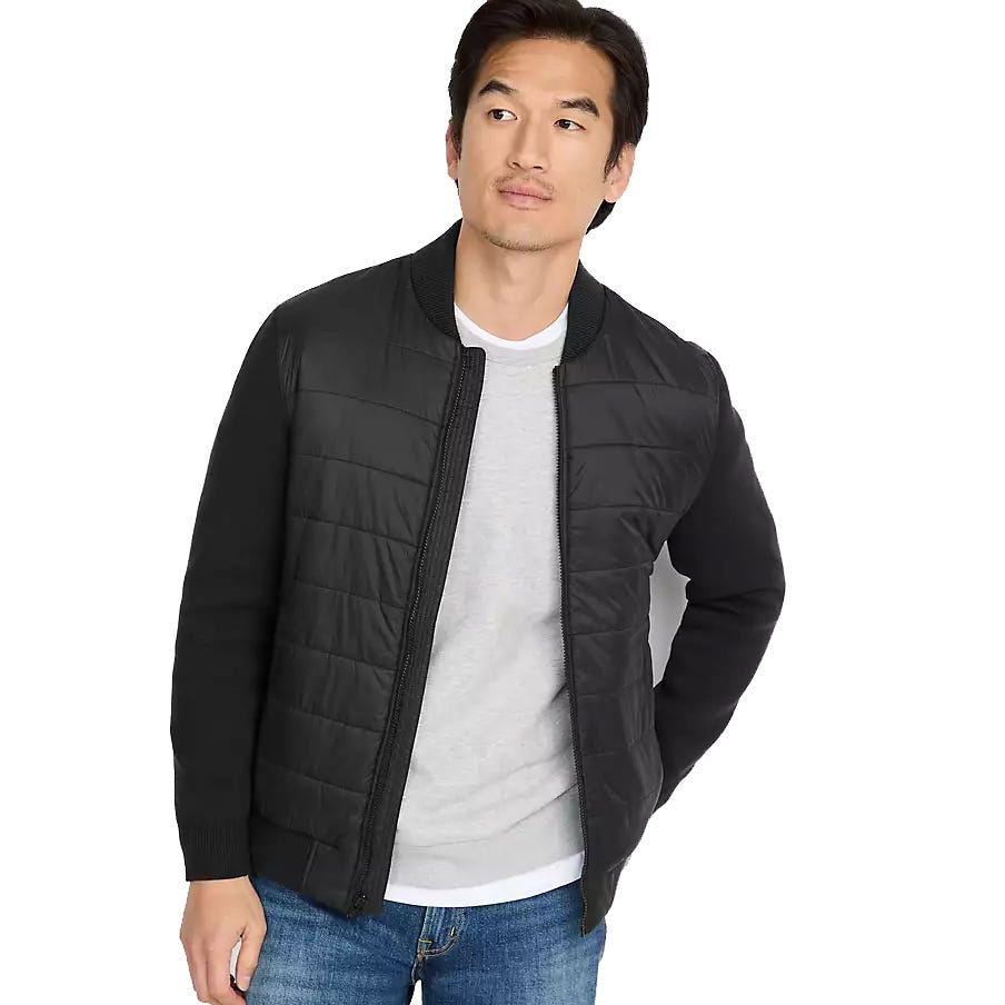 A man is wearing a black quilted bomber jacket over a gray sweater with blue jeans.
