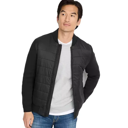A man is wearing a black quilted bomber jacket over a gray sweater with blue jeans.