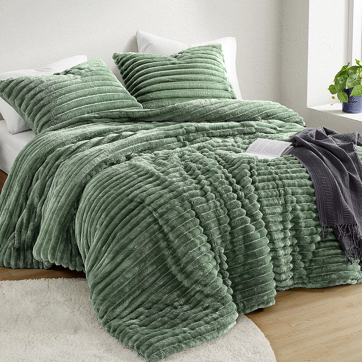 A textured green Queen Plush Comforter Set includes a ribbed comforter and matching pillow shams, creating a cozy and inviting bedroom aesthetic. A gray throw is draped over the bed, and a potted plant decorates the corner by the window.