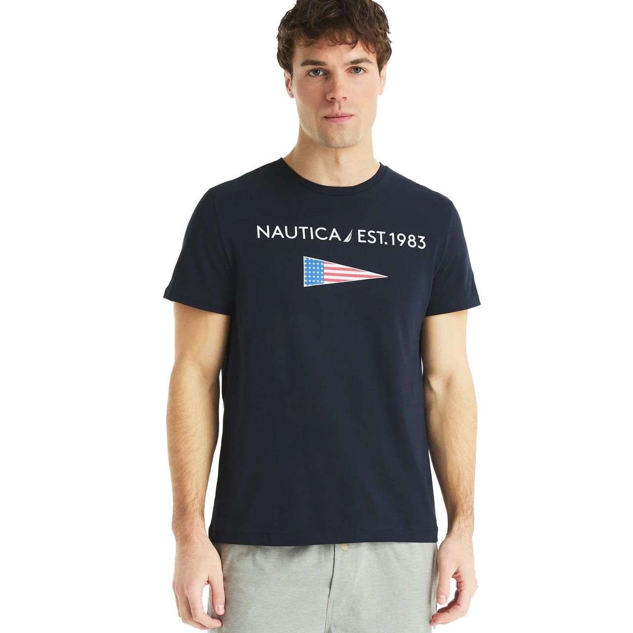 A man is wearing a navy blue Nautica T-shirt with \“NAUTICA EST. 1983\“ and a stylized American flag graphic.