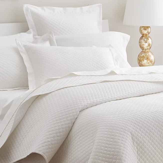 The 210TC Cotton Queen Coverlet Set features a white quilted coverlet with matching pillow shams, offering a textured design that adds a touch of elegance. A gold-toned lamp with a spherical base complements the bedding's clean and simple aesthetic.