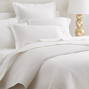 The 210TC Cotton Queen Coverlet Set features a white quilted coverlet with matching pillow shams, offering a textured design that adds a touch of elegance. A gold-toned lamp with a spherical base complements the bedding's clean and simple aesthetic.