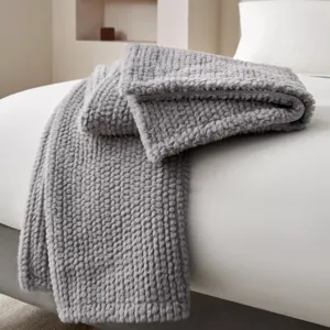 A soft, gray, textured knit blanket is draped over a white bed, adding a cozy touch to the minimalist bedroom setting.