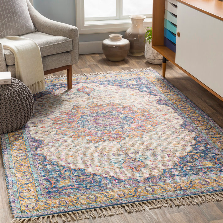 A 5' x 7' area rug with a vintage-inspired design features a pastel floral medallion pattern in blue, pink, and beige hues, finished with fringe edges, placed on a hardwood floor beside a gray armchair, knitted pouf, and vases near a window.