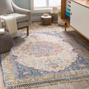 A 5' x 7' area rug with a vintage-inspired design features a pastel floral medallion pattern in blue, pink, and beige hues, finished with fringe edges, placed on a hardwood floor beside a gray armchair, knitted pouf, and vases near a window.