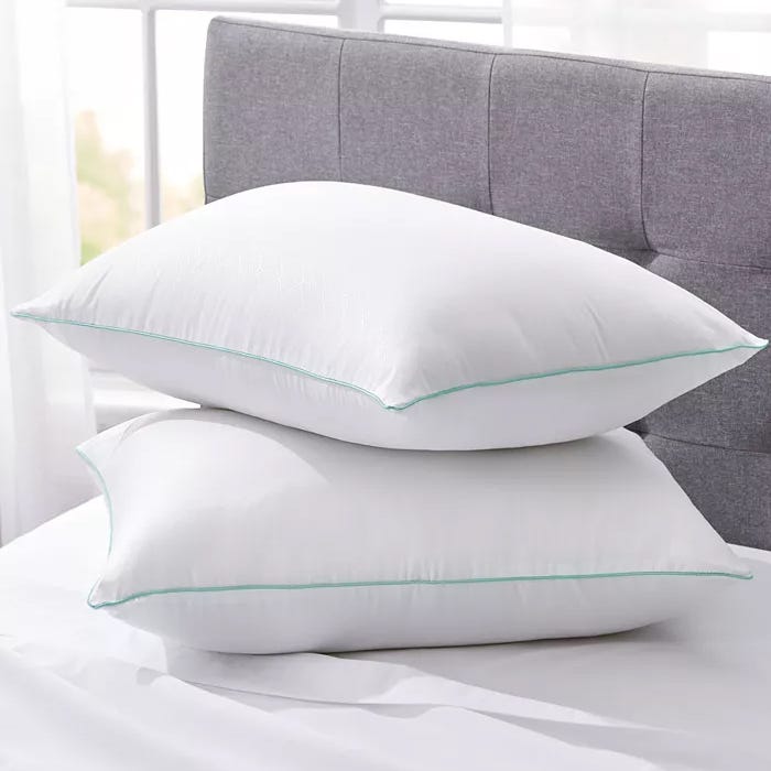 Two white pillows with light green piping are stacked on a bed with a gray headboard.