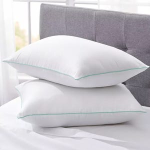 Two white pillows with light green piping are stacked on a bed with a gray headboard.