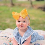 A baby wearing a blue and white striped outfit with a large yellow bow headband is sitting on a quilt.