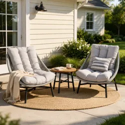 Outdoor patio setup with two cushioned lounge chairs and a small round table on a round woven rug, placed on a concrete surface beside a house with sliding glass doors and surrounded by greenery.