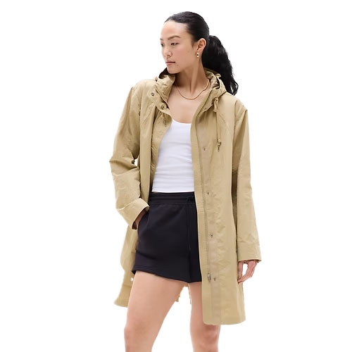 A person is wearing a beige long raincoat with a hood, over a white tank top and black shorts.