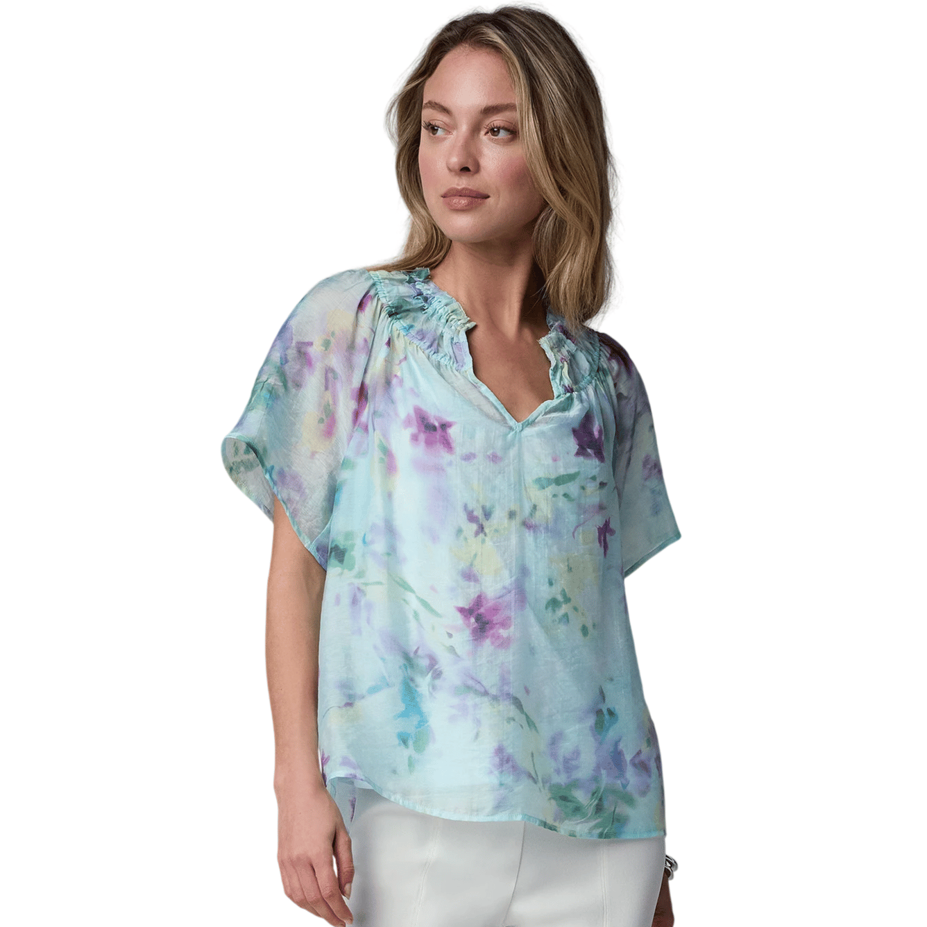 A woman is wearing a light, watercolor-patterned blouse with a ruffled neckline and short sleeves, paired with white pants.