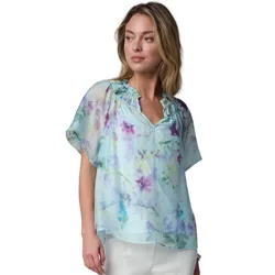 A woman is wearing a light, watercolor-patterned blouse with a ruffled neckline and short sleeves, paired with white pants.