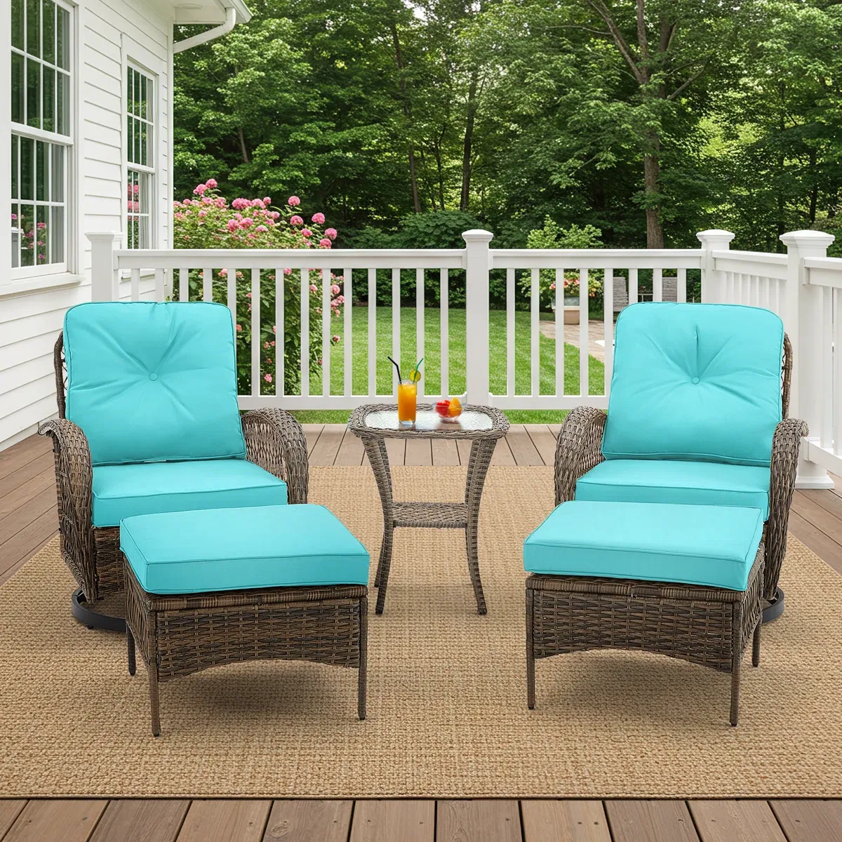 Two wicker chairs with turquoise cushions and matching ottomans are set on a wooden deck with a small round wicker table between them. The deck is bordered by a white railing, with greenery and pink flowers in the background.