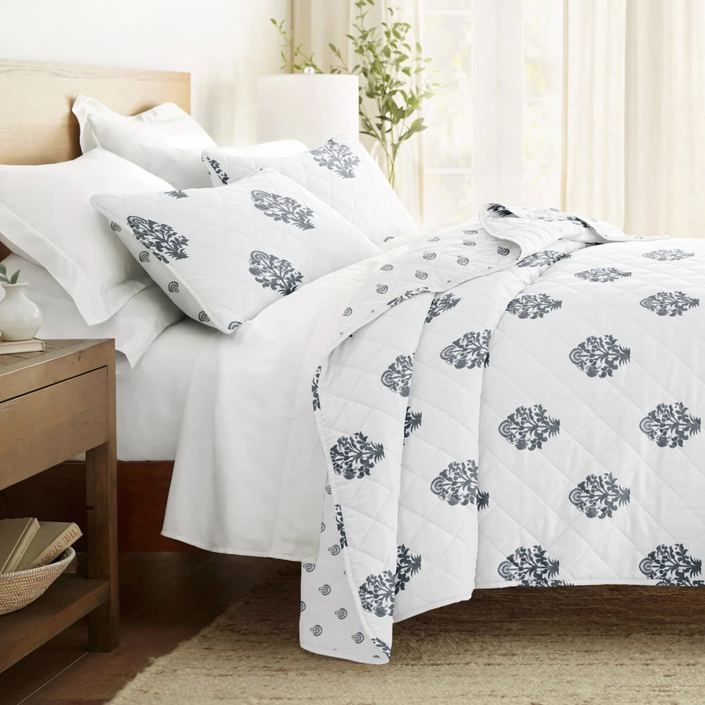 A neatly arranged bed features a white quilt and matching pillowcases with blue floral patterns, set against a warm and cozy room with wooden furniture and a potted plant.