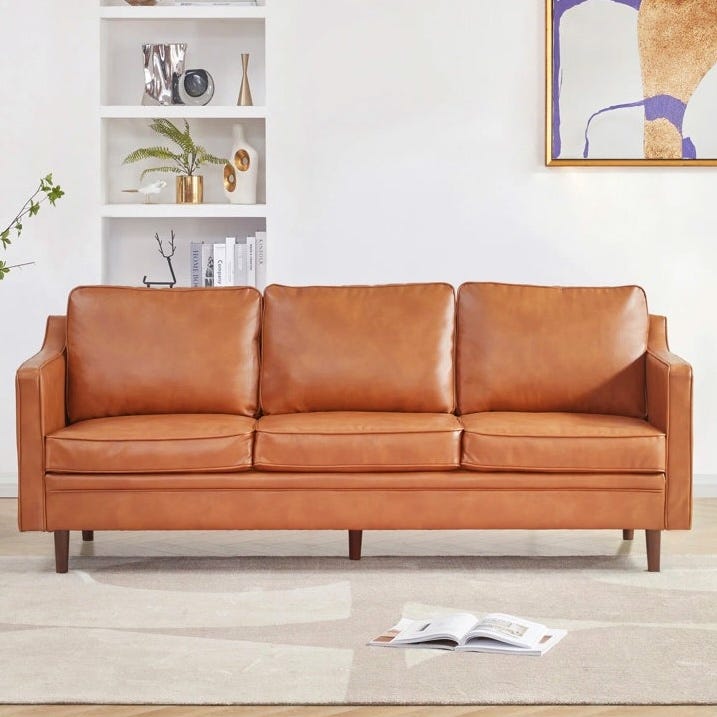A brown leather sofa with three cushions is set in a living room, featuring a minimalist design with wooden legs. The background includes white shelves, decorative items, and abstract wall art.