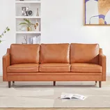 A brown leather sofa with three cushions is set in a living room, featuring a minimalist design with wooden legs. The background includes white shelves, decorative items, and abstract wall art.