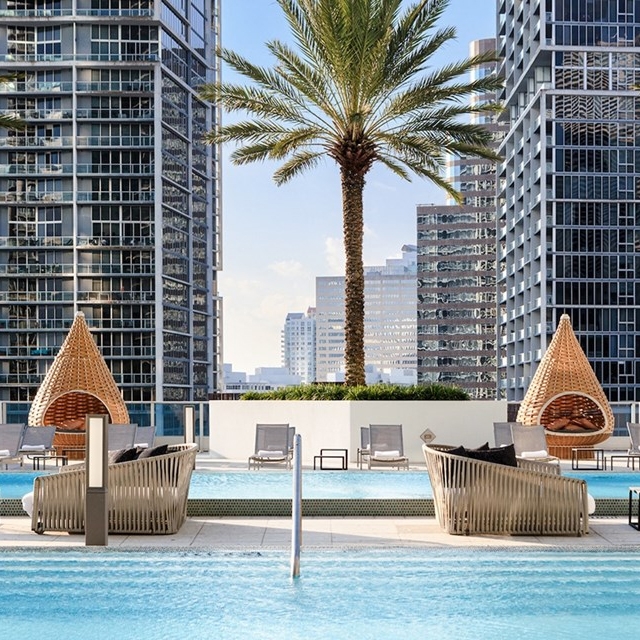 A rooftop pool area with wicker lounge chairs and egg-shaped cabanas, surrounded by tall modern buildings and a central palm tree.