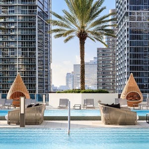 A rooftop pool area with wicker lounge chairs and egg-shaped cabanas, surrounded by tall modern buildings and a central palm tree.