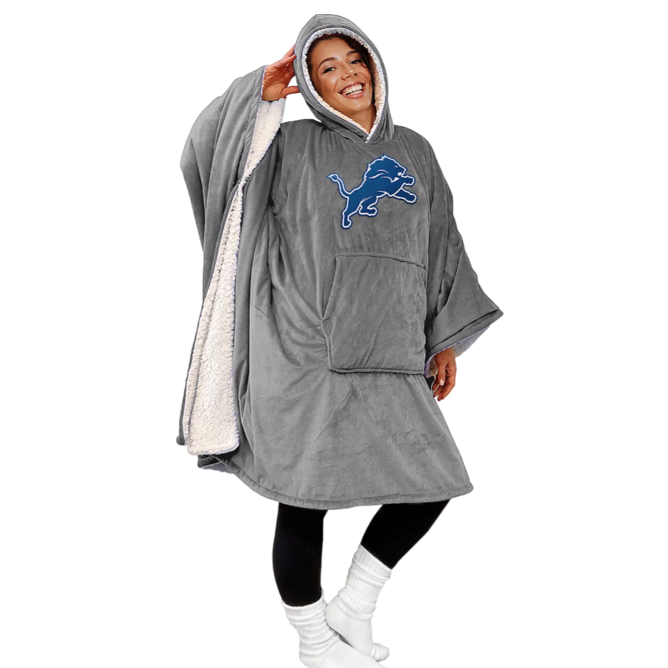 A person is wearing a cozy, oversized hooded blanket with a Detroit Lions logo. The blanket is gray with a sherpa lining, featuring wide sleeves, a front pocket, and black leggings paired with white socks.