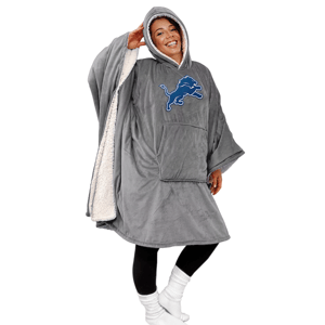 A person is wearing a cozy, oversized hooded blanket with a Detroit Lions logo. The blanket is gray with a sherpa lining, featuring wide sleeves, a front pocket, and black leggings paired with white socks.