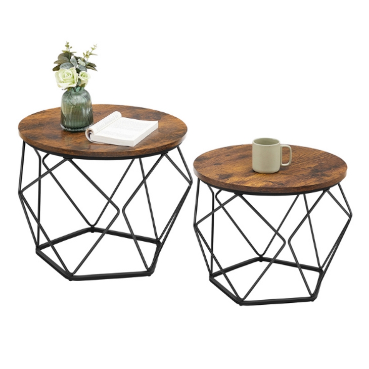 Two round side tables with rustic wooden tops and black metal geometric frames. One table holds a green vase with white flowers and an open book, while the other has a green mug.