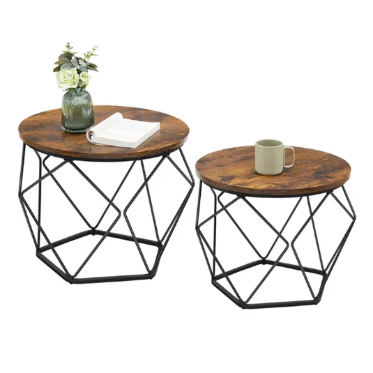 Two round side tables with rustic wooden tops and black metal geometric frames. One table holds a green vase with white flowers and an open book, while the other has a green mug.