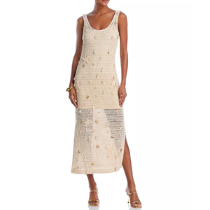 A beige crochet sleeveless maxi dress featuring a scoop neckline and embellished with small shells. It has a semi-transparent lower section with a side slit, paired with strappy high-heeled sandals.