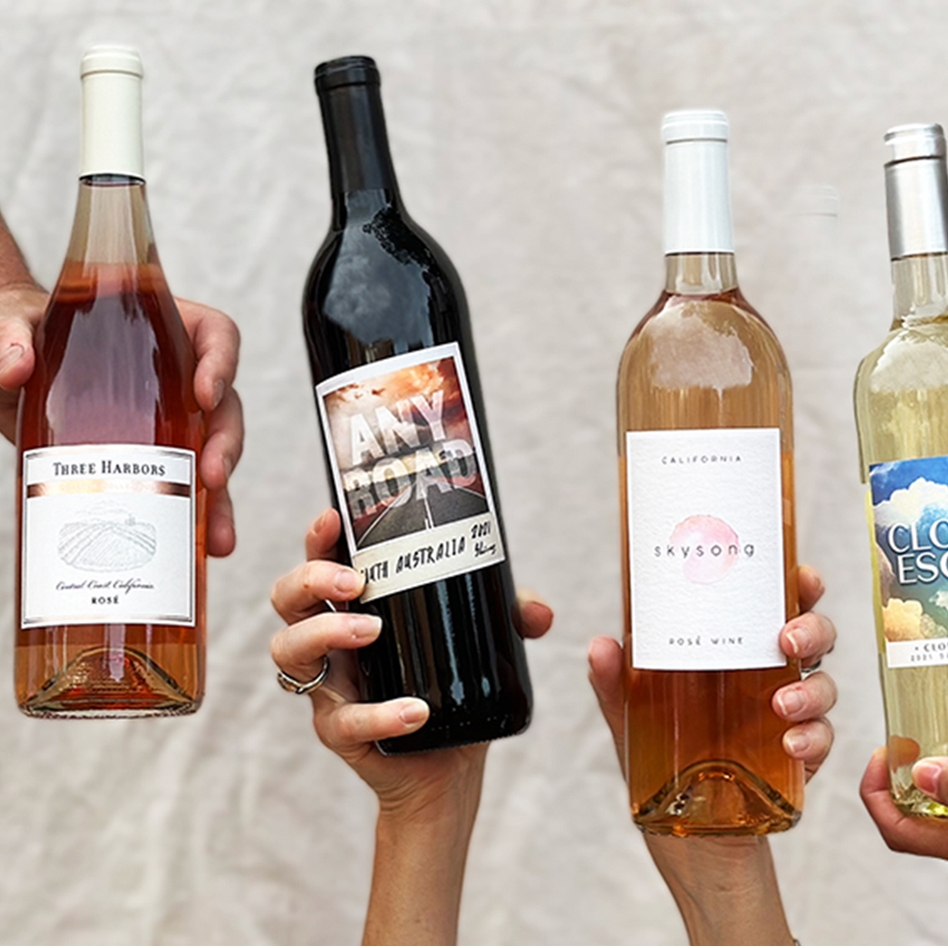 Four bottles of wine are being held by different hands. The labels read: \“Three Harbors\“ Rosé, \“Any Road\“ from South Australia, \“Skysong\“ California Rosé, and \“Cloud Escape\“ Chardonnay.