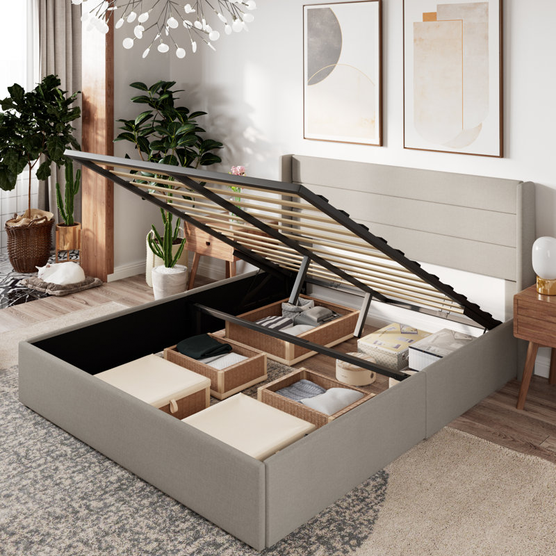 A lift-up bed with under-bed storage compartments is organized with baskets and boxes. The surrounding room has modern decor, with plants, abstract wall art, and a contemporary chandelier.