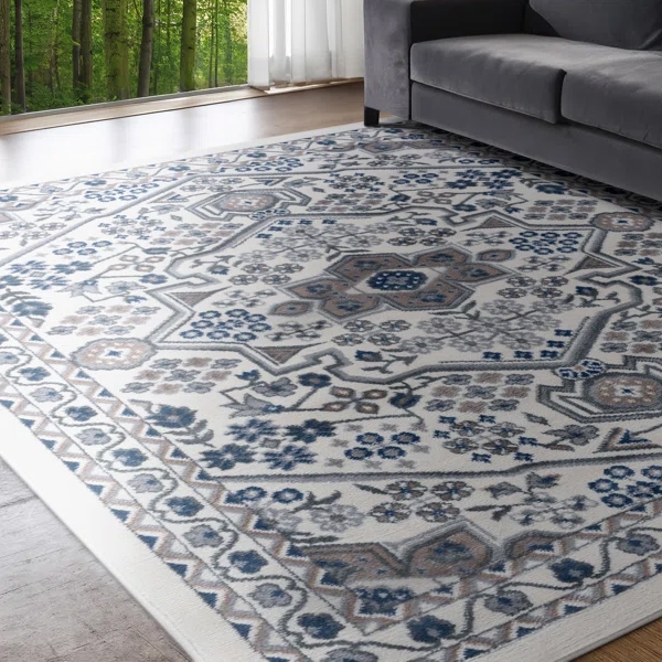 A patterned rug with a grey, blue, and beige floral motif lies on a wooden floor next to a gray couch, with a forest view through large windows in the background.