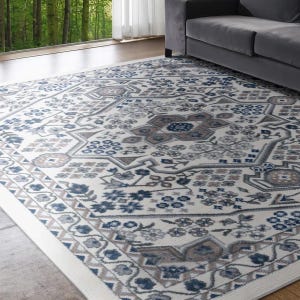 A patterned rug with a grey, blue, and beige floral motif lies on a wooden floor next to a gray couch, with a forest view through large windows in the background.