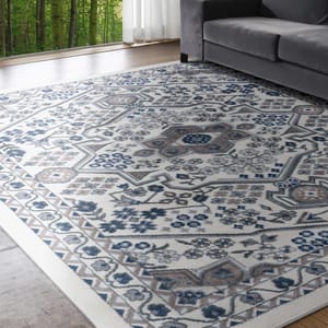 A patterned rug with a grey, blue, and beige floral motif lies on a wooden floor next to a gray couch, with a forest view through large windows in the background.