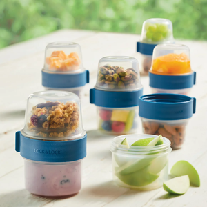 Stackable containers with blue lids contain various snacks like yogurt, granola, fruit, nuts, and apple slices.