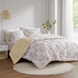 A bedroom features a floral-patterned comforter set with matching pillows, a bedside table with a small framed picture, and a vase with yellow flowers. A woven hanging lamp and sheer curtains complete the cozy and serene decor.